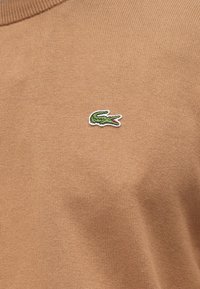 Rjavo pleten pulover z majhnim zelenim logotipom krokodila, izvezenim na levi strani. Tkanina je gladka in lahka.