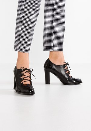Black patent leather high-heeled shoes with open lace-up design worn with cropped black-and-white houndstooth patterned pants.