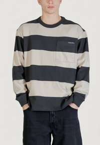 Maglia a maniche lunghe a righe in cotone beige e blu navy scuro, con tasca sul petto e collo a coste rotondo. Vestibilità ampia con maniche risvoltate.