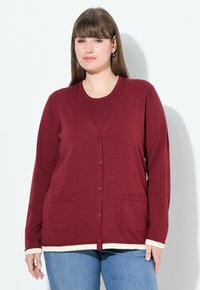 Kvinna med rakt brunt hår som bär gyllene ringörhängen, en bourgognyfärgad knäppt cardigan med fickor och ljusblå jeans mot en vit bakgrund.