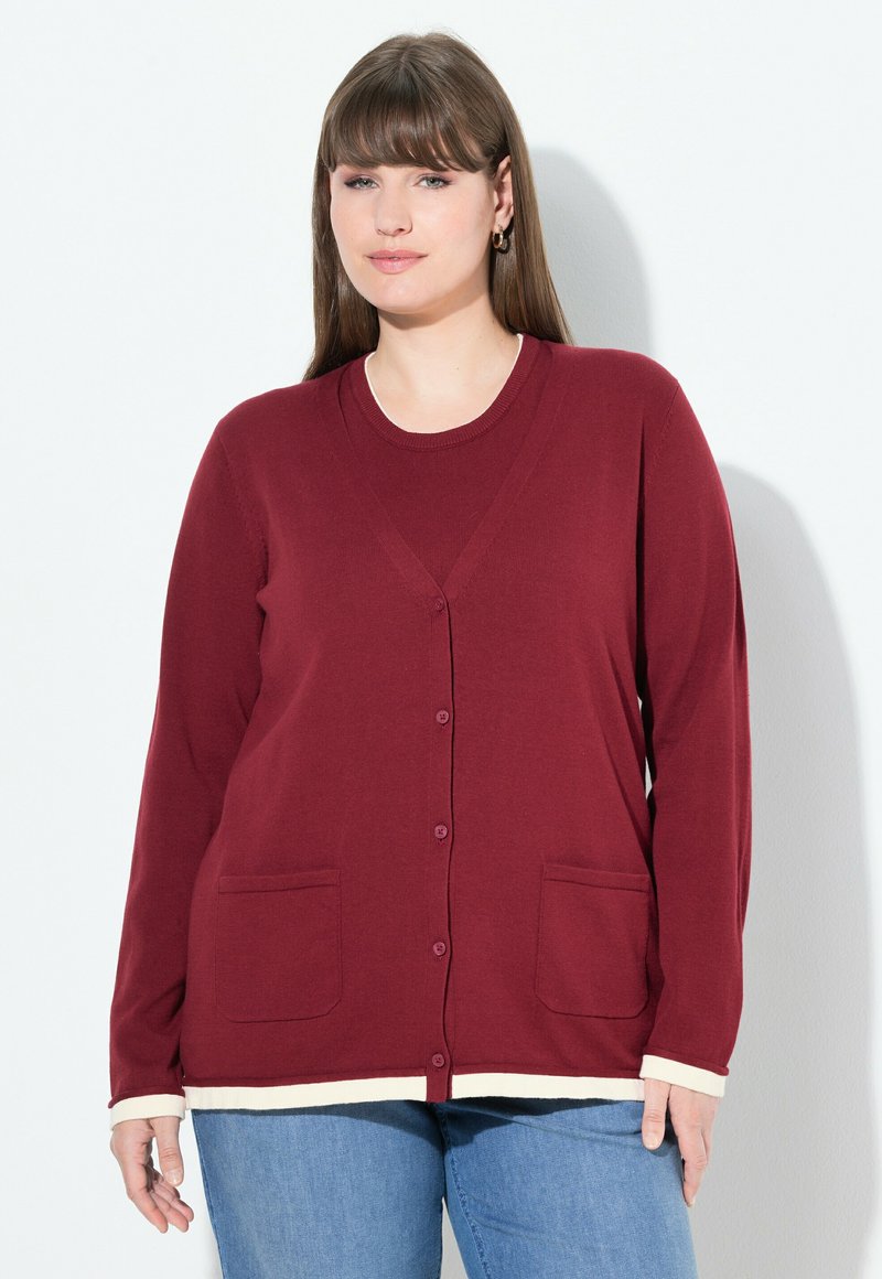 Kvinna med rakt brunt hår som bär gyllene ringörhängen, en bourgognyfärgad knäppt cardigan med fickor och ljusblå jeans mot en vit bakgrund.