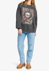 Sudadera gris con un gráfico de "Waves n' Rays" en colores suaves, combinada con jeans azul claro y botas marrones de suela gruesa.