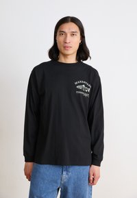 Black long-sleeve t-shirt made of cotton, featuring a printed graphic of a fish and text "MANASTASH FISHING CLUB" in white and green. Casual fit.