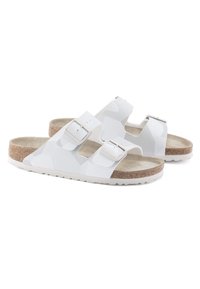 White slip-on sandals with two adjustable buckles, cork footbed, textured upper, and patterned design. Rubber sole provides traction.