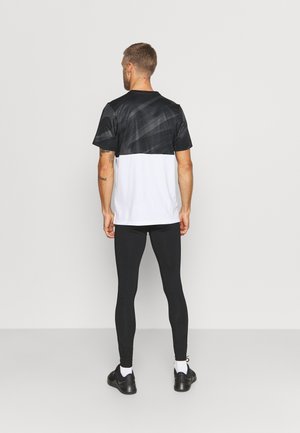 Mann mit schwarzem und weißem kurzärmeligem Shirt, schwarzen Leggings, weißen Socken und schwarzen Turnschuhen, steht mit dem Rücken zur Kamera vor schlichtem Hintergrund.