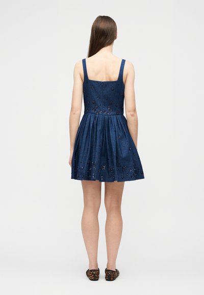 Γυναίκα με μακριά καστανά μαλλιά, φορώντας ένα χωρίς μανίκια μπλε φόρεμα με κεντήματα λουλουδιών και φλατ παπούτσια λεοπάρδαλης, στέκεται γυρισμένη προς τα πίσω πάνω σε λευκό φόντο.