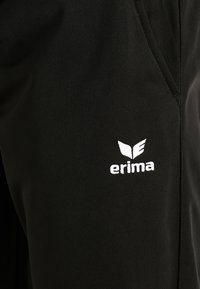 Pantalons de sport noirs en tissu lisse, arborant un logo "erima" blanc et un design d'aile distinctif. Style simple et fonctionnel.