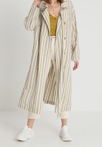Pruhovaný, oversize trenčkot v krémové barvě s modrými pruhy; má velké knoflíky, široké rukávy a splývavý střih. Nosený přes hořčicový top.