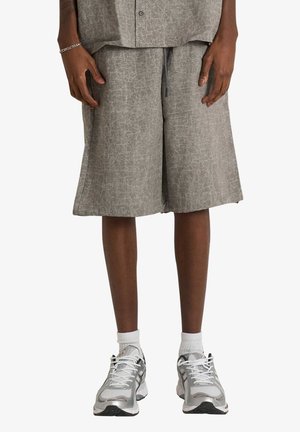 Persona che indossa pantaloncini larghi grigi testurizzati fino al ginocchio, calzini bianchi e scarpe da ginnastica argento e bianche, in piedi con le mani lungo i fianchi.