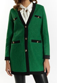 Cárdigan verde con textura, con acentos negros, botones dorados, cuello con muesca y bolsillos delanteros, combinado con una blusa blanca y pantalones negros.