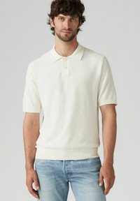 Kortärmad, kragad pikétröja i ljus krämfärg, i strukturerad stickad kvalitet. Har två knappar vid kragen samt ribbad fåll och ärmslut.