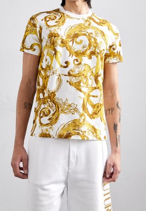 Mand iført en hvid kortærmet t-shirt med guldornate barokmønstre og hvide bukser med guldtekst på siden.