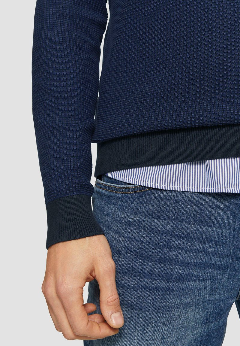 Marinefarbener, strukturierteter Pullover mit gerippten Bündchen, über einem blau-weiß gestreiften Hemd getragen, kombiniert mit Jeans. Nahaufnahme von Hand- und Stoffdetails.