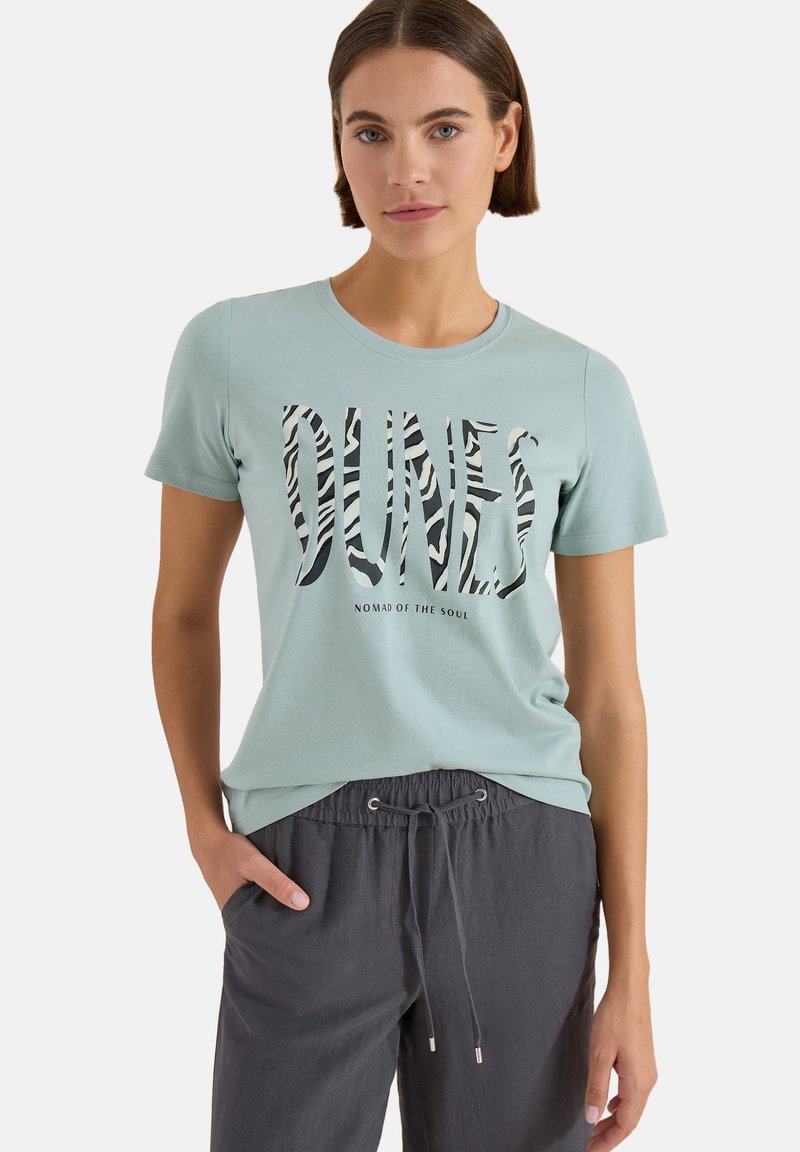 Femme portant un t-shirt bleu clair avec "DUNES" en imprimé zèbre et le texte "NOMAD OF THE SOUL", associé à un pantalon gris foncé avec cordon.