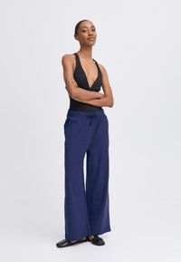 Svart halterneck bodysuit i kombination med lösa, marinblå vida byxor med dragsko i midjan och platta sandaler. Enkla linjer och andningsbart tyg.