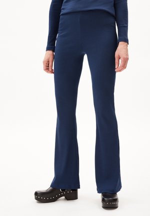 Persoon draagt marineblauwe geribbelde uitlopende broek en zwarte gestudeerde platformklompen, staand tegen een effen witte achtergrond.