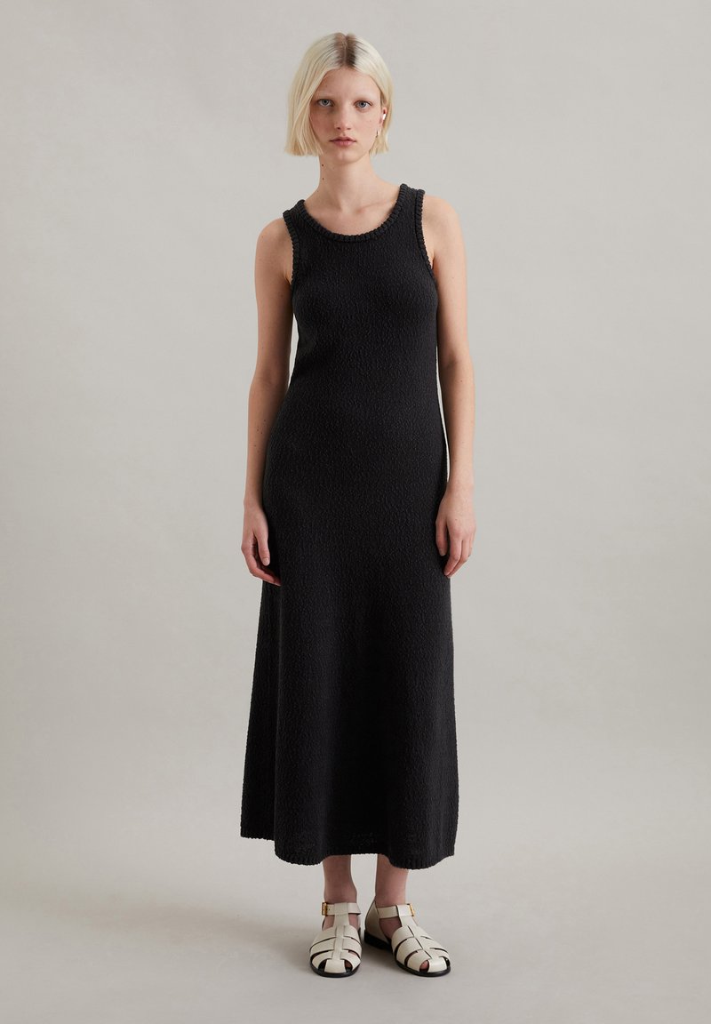 Zwarte mouwloze maxi-jurk van textuurstof met een ronde halslijn. Model draagt witte sandalen met banden en gouden accenten.