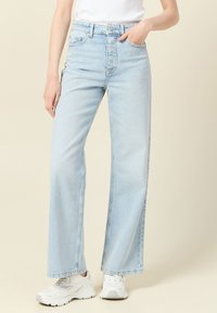 Jeans de tiro alto de pierna ancha de color azul claro con dobladillo deshilachado, cinco bolsillos y cierre de botón. Combinados con zapatillas deportivas blancas.