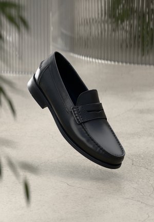Mocassino da uomo nero in pelle con cucitura moc toe e fascia decorativa sulla tomaia, mostrato sospeso sopra una superficie chiara e strutturata.
