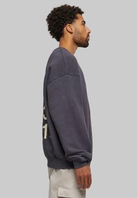 Sudadera gris oscuro de manga larga, con puños acanalados y detalles de costura sutiles. Presenta texto en dorado en un lateral.