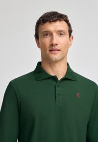 Dunkelgrünes Langarm-Polohemd mit Kragen, mit einem roten gestickten Logo auf der linken Brust und einer dreiknöpfigen Vorderseite.