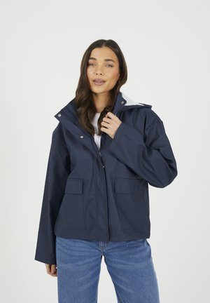 Marineblauwe waterdichte jas met capuchon, ritssluiting, drukknopen en twee voorkeuzes; gedragen over een witte top en blauwe spijkerbroek.