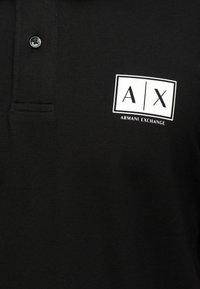 Μαύρο πόλο μπλουζάκι με λευκό ορθογώνιο λογότυπο Armani Exchange που απεικονίζει "A | X" στο στήθος κοντά στα κουμπιά.