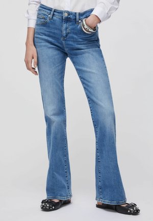 Personne portant un jean évasé bleu clair avec la main dans la poche, associée à des chaussures slip-on noires cloutées sur un fond gris.