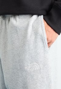 Kišenėje laikomą ranką, vilkint lengvus šviesiai pilkus sportinius kelnes su „The North Face“ logotipu, suderintus su juoda ilgomis rankovėmis marškiniais.