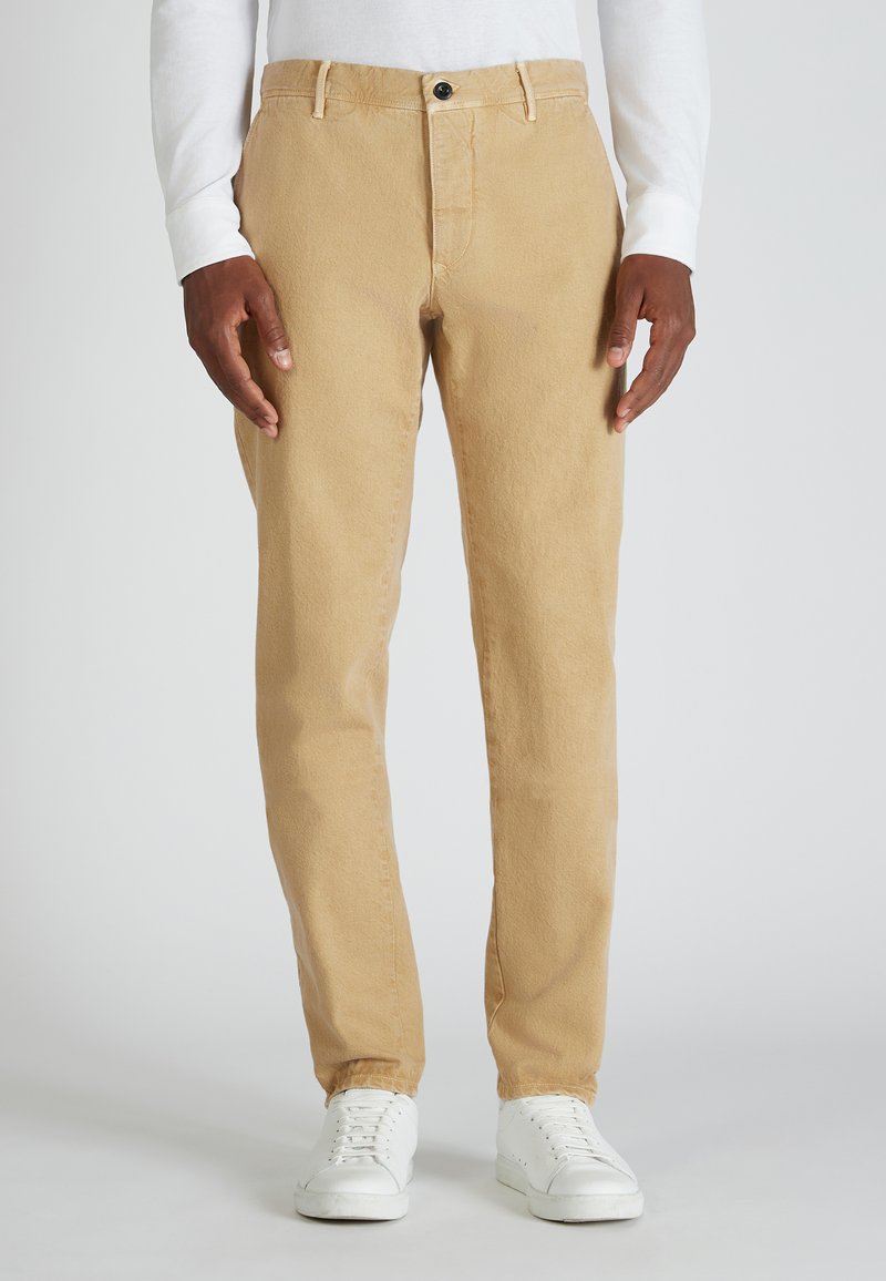 Hombre con pantalones rectos beige, camisa de manga larga blanca y zapatillas blancas, de pie contra un fondo gris liso.