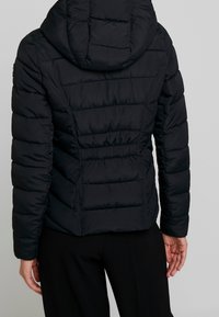 Sort pufferjakke med hætte, der har vandrette quiltede paneler og slim fit talje. Materialet ser glat og isoleret ud.
