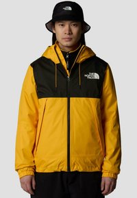 Gelbe und schwarze wasserdichte Jacke mit einem Frontreißverschluss, verstellbare Kapuze und elastischen Bündchen. Begleitet von einem schwarzen Bucket Hat.