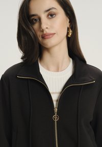 Giacca nera con zip e colletto alto, dotata di dettagli in oro, indossata sopra una maglietta bianca con texture e righe orizzontali.