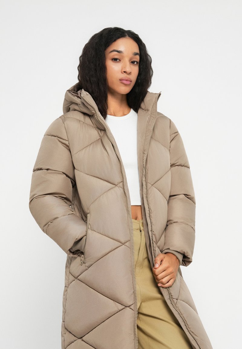 Femme aux longs cheveux bruns portant un manteau à capuche beige matelassé, un haut blanc court et un pantalon kaki sur fond blanc uni.