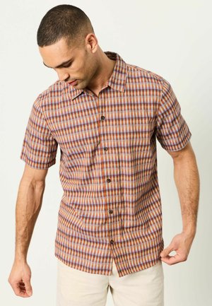 Mann mit kurzen Haaren, der ein kurzärmeliges Hemd mit orange-blau-braunem geometrischem Muster trägt und den Saum über einer hellen Hose richtet.