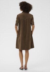 Brązowa sukienka koszulowa z dżerseju z krótkimi rękawami, plisowanym tyłem i bocznymi kieszeniami. Długość dołem kończy się powyżej kolana.
