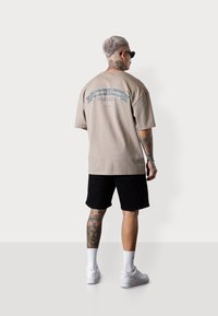 T-shirt en coton beige avec un design graphique dans le dos, associé à un short noir et des baskets blanches. Tatouages visibles sur la jambe inférieure.
