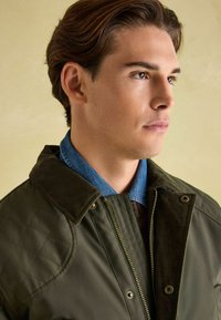 Wypchana oliwkowa kurtka z kołnierzem z frotte, kontrastująca z niebieską koszulą z denimu pod spodem i fakturowaną brązową warstwą wełnianą.