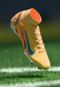 Scarpetta da calcio Nike di un giallo acceso con accenti blu, tomaia testurizzata, interno arancione e tacchetti multicolore, sospesa sopra l'erba verde.