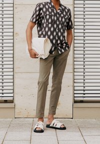 Camicia a maniche corte bianca e nera a fantasia, pantaloni slim-fit verde oliva, borsa a tracolla crema e sandali bianchi con suole nere.