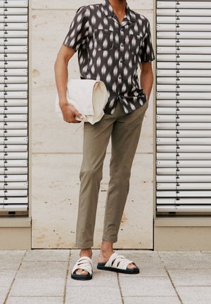 Camicia a maniche corte bianca e nera a fantasia, pantaloni slim-fit verde oliva, borsa a tracolla crema e sandali bianchi con suole nere.