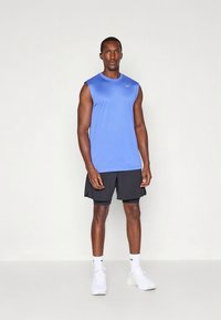 Blå ärmlös sporttopp för män med rund halsringning och logotyp, kombinerad med svarta shorts. Outfiten fullbordas med vita sportskor.