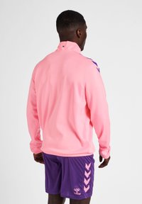 Giacca sportiva rosa con colletto alto e accenti viola sulle spalle, abbinata a pantaloni corti viola con motivo a zigzag rosa sui lati.