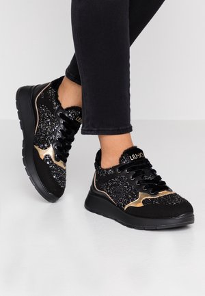 Baskets noires avec des paillettes et des accents dorés, lacets noirs et logo de la marque, portées avec un pantalon noir ajusté sur un sol clair.