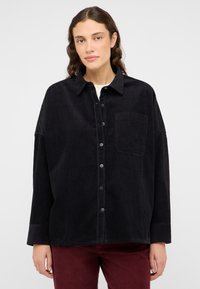 Camisa de pana negra con cuello, cierre de botones en la parte delantera y un bolsillo en el pecho. Presenta mangas anchas y una textura acanalada.