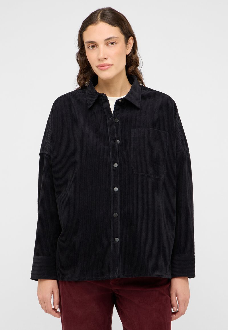 Camisa de pana negra con cuello, cierre de botones en la parte delantera y un bolsillo en el pecho. Presenta mangas anchas y una textura acanalada.