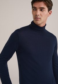Joven con un suéter de cuello alto azul marino mirando hacia su izquierda frente a un fondo beige liso.