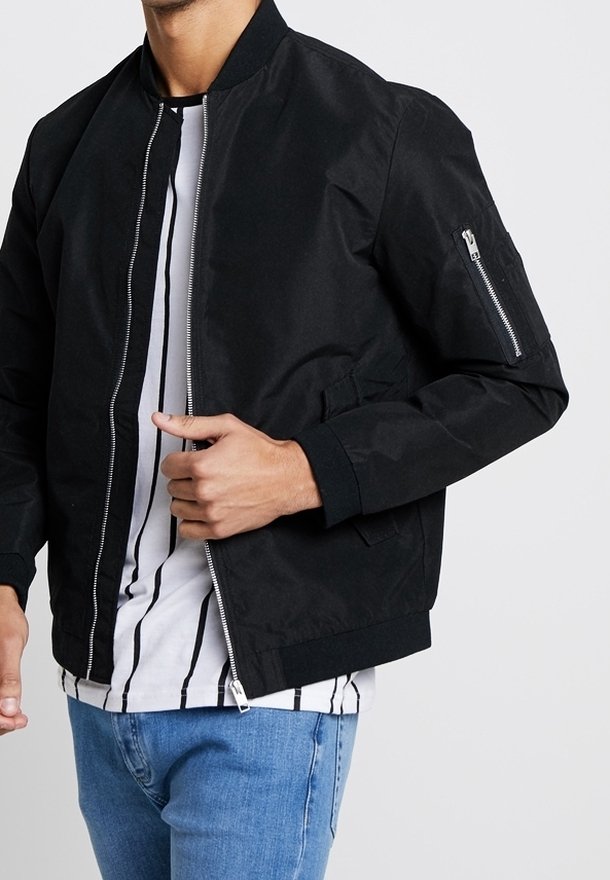 Homme portant une veste bomber noire par-dessus une chemise rayée blanche et noire avec un jean bleu, debout devant un fond uni.