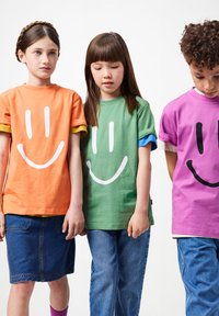 Tres niños están de pie uno al lado del otro, vistiendo camisetas de caras sonrientes coloridas y jeans, mirando hacia abajo o de frente contra un fondo blanco.