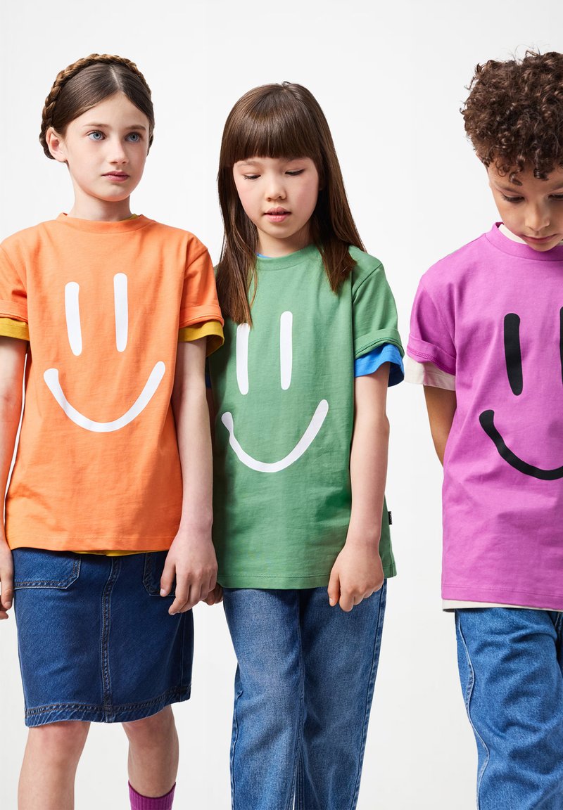 Tres niños están de pie uno al lado del otro, vistiendo camisetas de caras sonrientes coloridas y jeans, mirando hacia abajo o de frente contra un fondo blanco.
